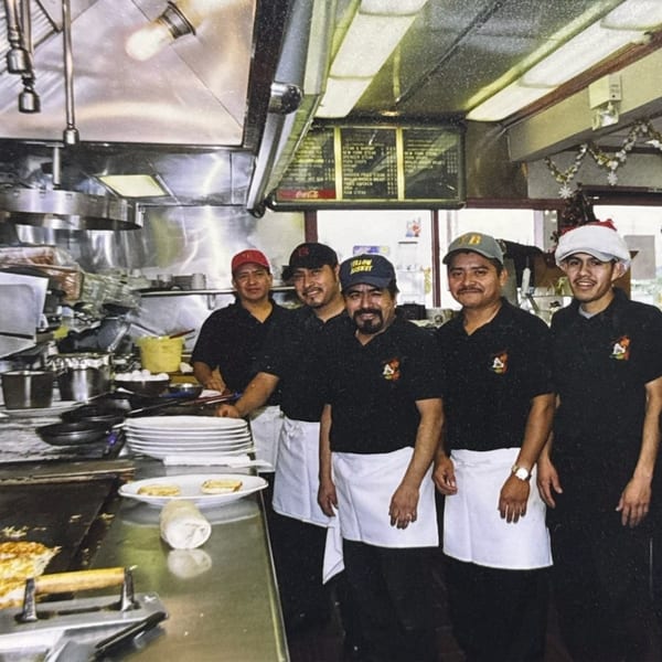 Six male kitchen staff in black shirts and white aprons smiling in a busy restaurant kitchen.