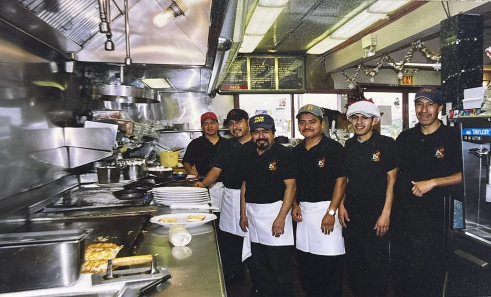 Six male kitchen staff in black shirts and white aprons smiling in a busy restaurant kitchen.