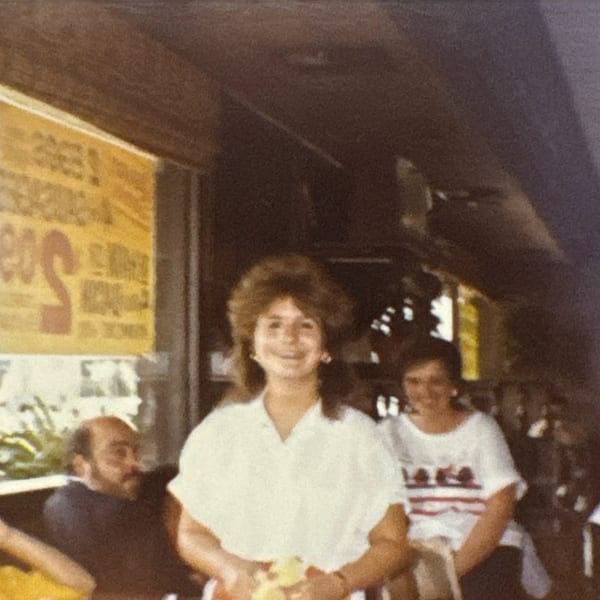 Smiling woman with big hair in a white shirt, with other patrons in a restaurant.