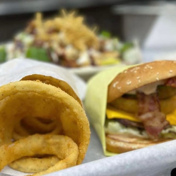 Crispy onion rings in a paper bag next to a bacon cheeseburger with a salad in the background.