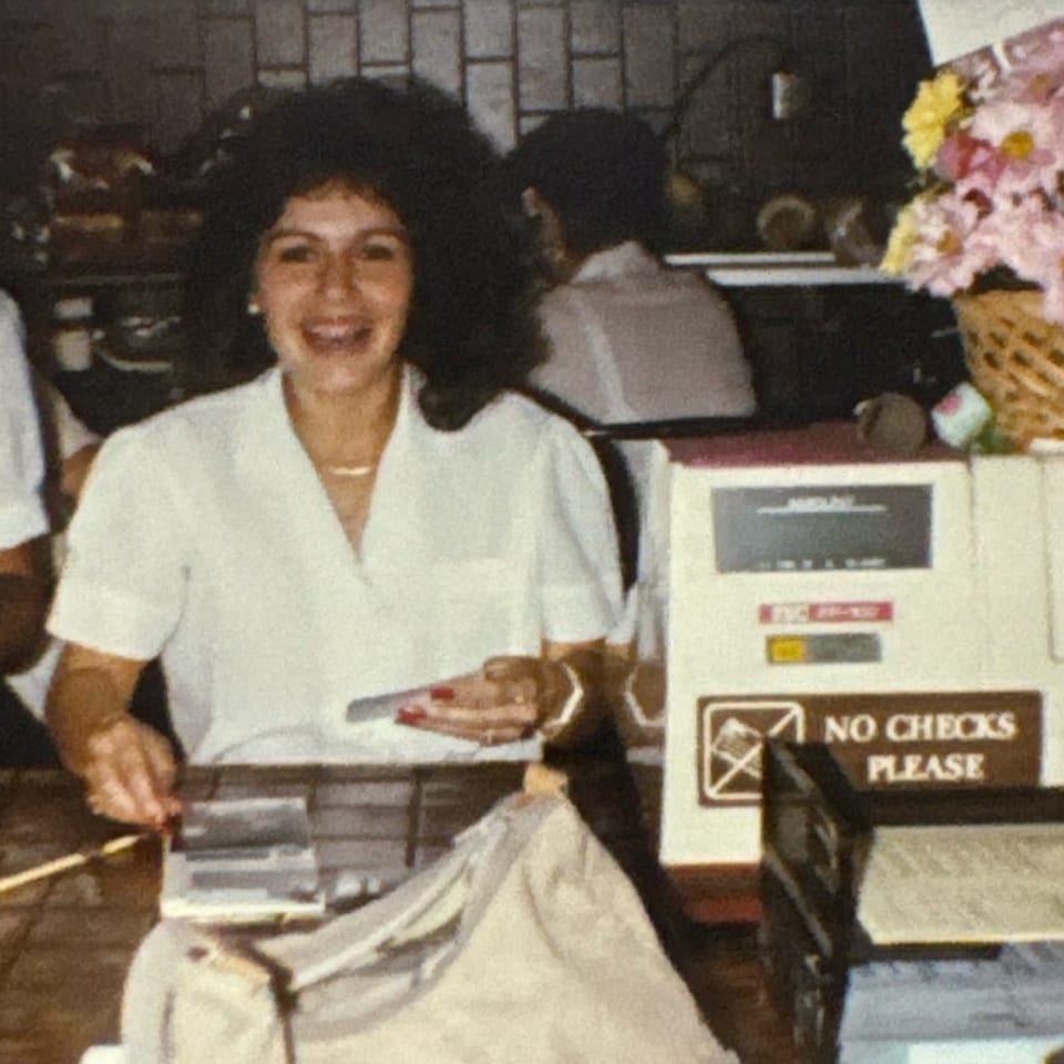 A smiling woman with curly hair works at a counter with a "No Checks Please" sign.