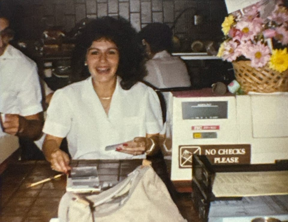 A smiling woman with curly hair works at a counter with a "No Checks Please" sign.