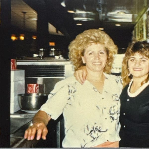Two women smiling, one with an arm around the other, in a restaurant kitchen with a chef.