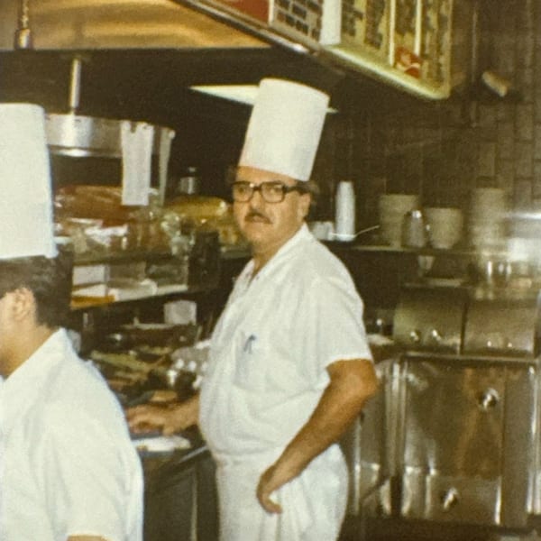 A chef in a tall white hat and glasses stands in a busy restaurant kitchen.