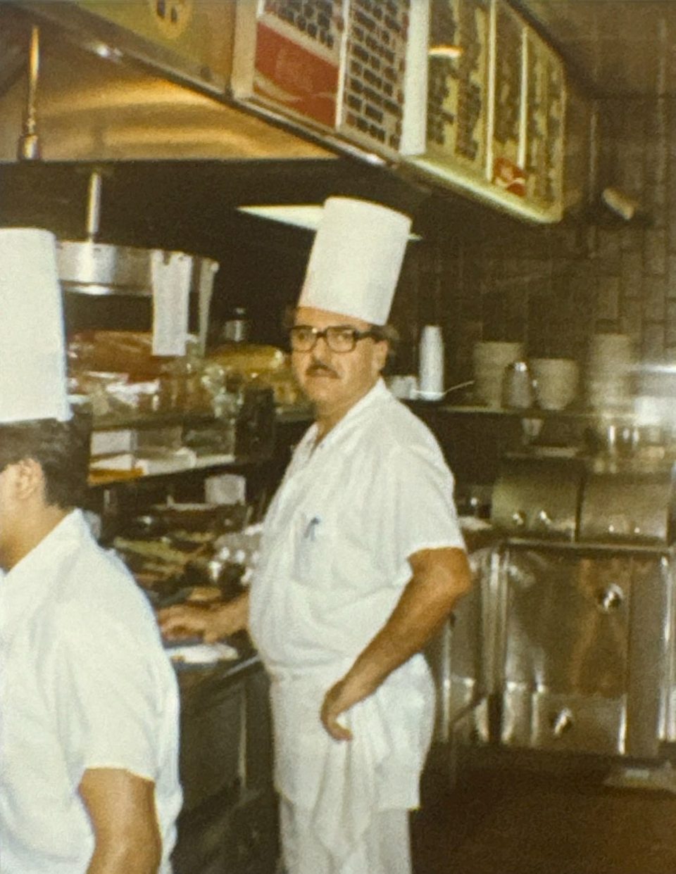 A chef in a tall white hat and glasses stands in a busy restaurant kitchen.