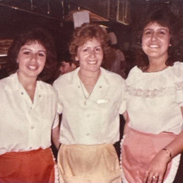 Three smiling women in white shirts and aprons stand together in a restaurant kitchen.
