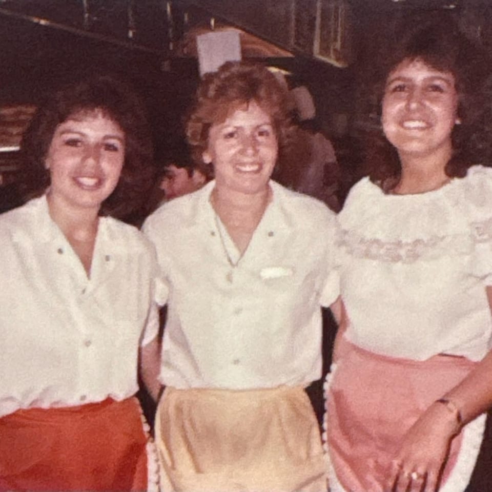 Three smiling women in white shirts and aprons stand together in a restaurant kitchen.