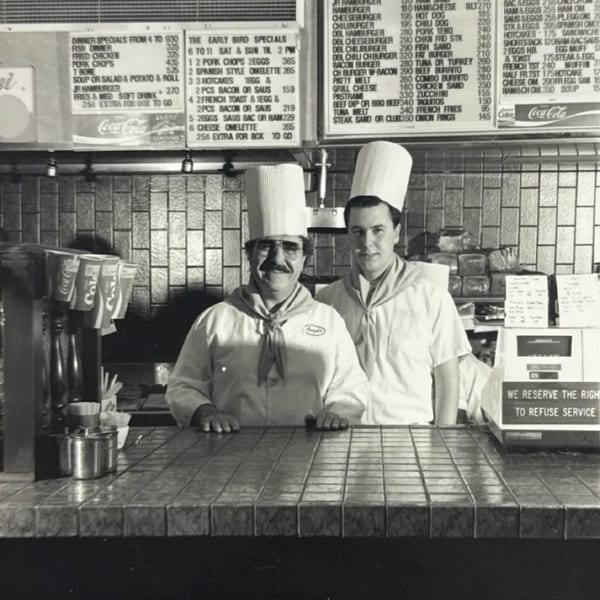 Two chefs in tall white hats and uniforms stand behind a tiled counter with menus above.
