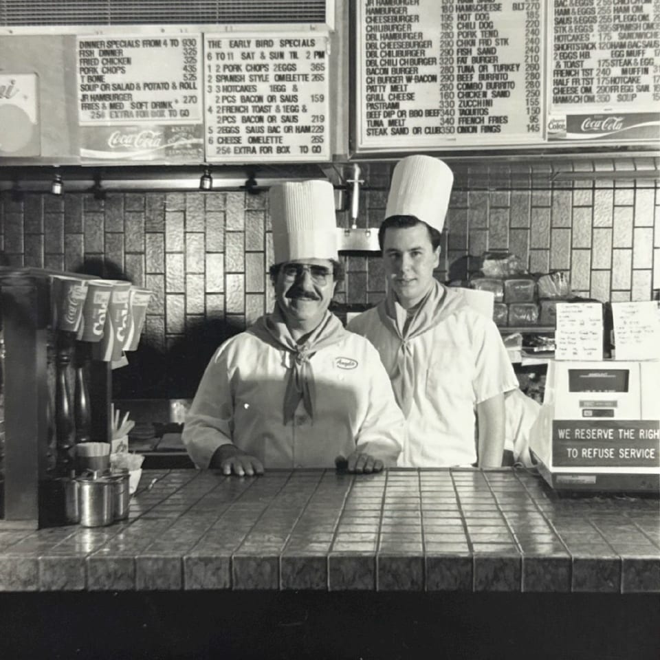 Two chefs in tall white hats and uniforms stand behind a tiled counter with menus above.