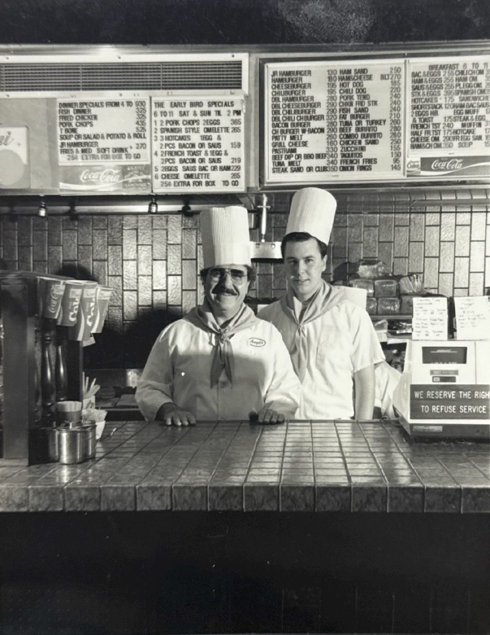 Two chefs in tall white hats and uniforms stand behind a tiled counter with menus above.