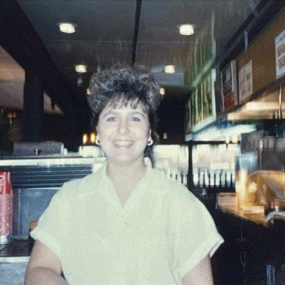Smiling woman with curly hair in a diner, with soda dispensers and menu boards in the background.