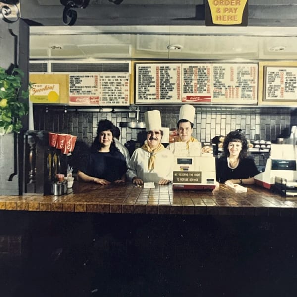 Four smiling restaurant staff members, two in chef hats, stand behind a counter with menus above.