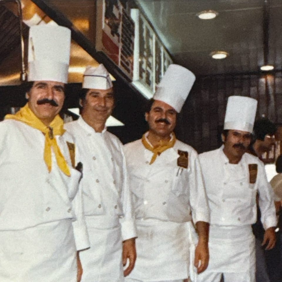 Four chefs in white uniforms and hats, two with yellow scarves, stand smiling in a kitchen.