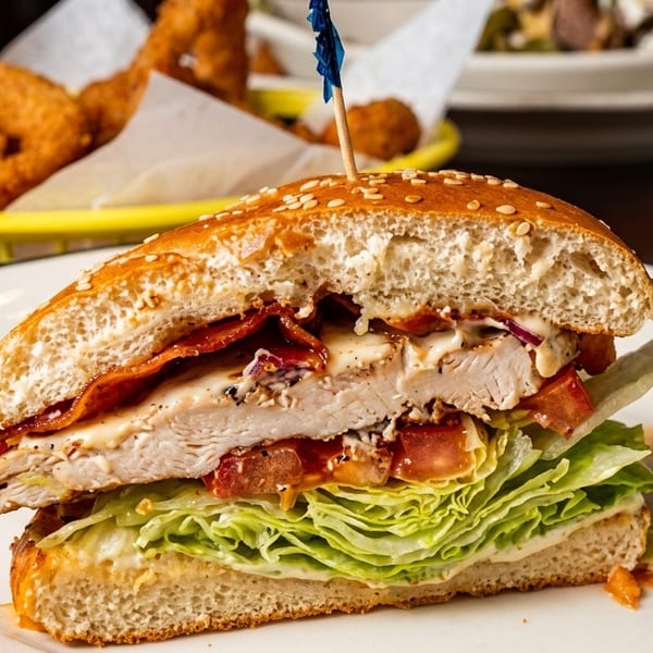 Chicken sandwich with bacon, lettuce, tomato, and sauce, with onion rings in the background.