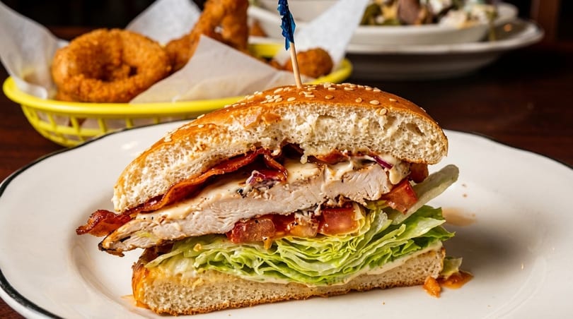 A chicken sandwich with bacon, lettuce, and tomato on a sesame bun, with onion rings in the background.